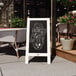 A white Flash Furniture A-Frame chalkboard sign on a sidewalk with a plant in a pot.