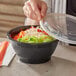 A person holding a Choice black plastic bowl with a lid over noodles and vegetables.