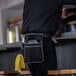 A black Ergodyne Arsenal server apron pouch with multiple pockets, attached to a person's waist and holding various items including a yellow cloth.