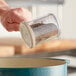 A hand holding a Carlisle shaker pouring ground black and brown spices into a pot.