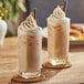 A close up of two glasses of Capora Original Chai Tea Lattes with whipped cream and cinnamon on a table.