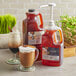 A plastic jug of Capora Original Chai Tea Latte concentrate on a table next to a glass of brown liquid.