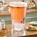 A large clear acrylic beverage dispenser with a spigot and an ice chamber at the bottom, filled with a pink drink and fruit slices.
