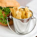 A person's hand holding a mini stainless steel pail of french fries with a sandwich on a table.