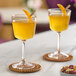 Two Arcoroc Romeo cocktail glasses filled with yellow liquid and topped with an orange slice on a table with a bowl of nuts and dried fruit.