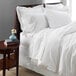 A white bed with a 1888 Mills Oasis white bed skirt on a bed with white sheets and a vase of flowers on a table.