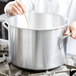 A person stirring food in a large silver Vollrath sauce pot with a wooden spoon.