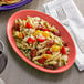 A Tuxton Cinnebar oval china coupe platter with pasta salad, tomatoes, and cheese with a fork on the table.