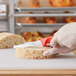 A hand with a red-handled Choice utility knife cutting bread on a cutting board.