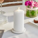 A white Hollowick wax pillar candle on a table with plates and glasses.