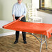 A woman rolling a tangerine plastic table cover onto a table outdoors.