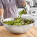 A woman pouring cucumbers into a Choice stainless steel mixing bowl on a wood surface.