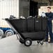 A man in a black shirt and pants pushing a black Lavex trash cart with garbage bags.