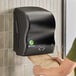 A black mechanical hands-free paper towel dispenser mounted on a tiled wall.