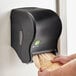 A person's hand putting a brown roll of paper towel in a black San Jamar Tear-N-Dry Essence EcoLogic paper towel dispenser.