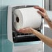 A person loading a large white hardwound paper towel roll into a wall-mounted dispenser.