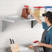 A man in a black apron and hat standing on a Regency stainless steel wall shelf holding a container of black and red food.