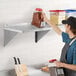 A man in a black apron and hat standing on a Regency stainless steel wall shelf holding containers of food and ground coffee.