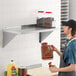 A man in a professional kitchen using a Regency stainless steel wall shelf to store plastic containers of seasonings.