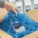 A hand holding a cardboard box filled with Spring-Fill Sky Blue Crinkle Cut Paper Shred.