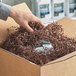 A hand holding a box of Spring-Fill Chocolate Crinkle Cut paper shred.