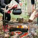 A man and woman cooking food over a BREEO stainless steel grill above a fire.