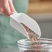 A close-up of a hand holding a white Vikan hand scoop full of seeds over a white container.