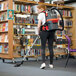 A woman wearing a vest using a NaceCare backpack vacuum to clean a book shelf.