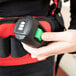A person's hand holding a green button to operate a NaceCare Solutions corded backpack vacuum with a battery power brush.