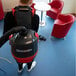 A person wearing a backpack vacuuming a white table with a metal base.