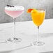 Two glasses of liquid, one orange and one pink, on a table with a Della Luce Aurora coupe glass.