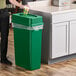 A person putting a cup in a Lavex dark green slim rectangular trash can with a Drop Shot lid.