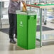 A woman in a green uniform putting food in a Lavex lime green rectangular trash can.