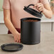 A hand holding a black Planetary Design Airscape container lid over a container of coffee beans.