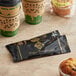 A black and gold Double Serving Hot Beverage Condiment packet next to a couple of coffee cups on a counter.