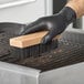 A person wearing black gloves using a Choice steel bristle grill brush head to clean a metal grill.