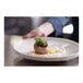 A person's hand holding a Libbey white porcelain plate with a piece of meat, cranberry sauce, and microgreens.