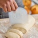 A person using a translucent plastic dough scraper to cut pieces from a loaf of dough on a floured surface.
