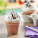 A clear plastic cup of chocolate milkshake with whipped cream and chocolate syrup next to another cup.