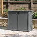 A Suncast grey plastic outdoor storage cabinet with a lid on a stone patio.