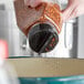 A person pouring seasoning into a pot using a black spice lid with 3 holes.