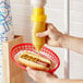 A person using an Asept hanging condiment pump to dispense mustard onto a hot dog.