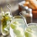 A close-up of a drink with lime and cucumber slices on a stainless steel skewer.