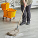 A person mopping the floor with a Lavex natural cotton wet mop on a wooden handle with a wire clamp.