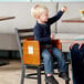 A wooden booster seat with a walnut finish designed for children, shown attached to a dining chair.