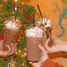 A group of three plastic cups filled with iced chocolate drinks, each topped with whipped cream and a black straw, held up in front of a decorated Christmas tree.