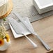 An Acopa Edgeworth stainless steel cake knife and fork on a white plate on a table with flowers.