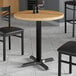 A Lancaster Table & Seating round MDF table top with a maple finish on a table with black chairs.