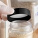 A person wearing gloves holding a plastic container with a black 63/485 polypropylene spice cap on it.