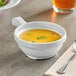 A white Acopa melamine soup bowl with a handle full of soup on a table with a spoon and fork.